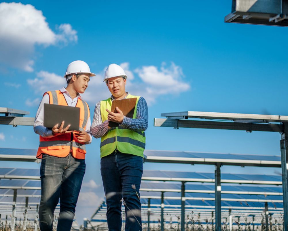 Young Asian technician man and colleague in safety uniform checking operation of sun and photovoltaic solar panel and use laptop computer while working in solar farm